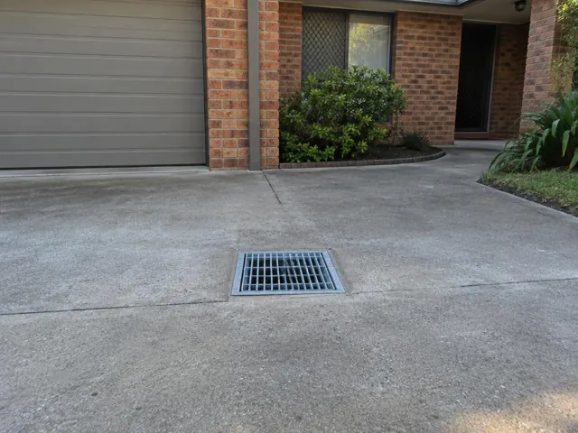 Concrete driveway with a clean outdoor drain grate beside brick walls and garden in Woodberry