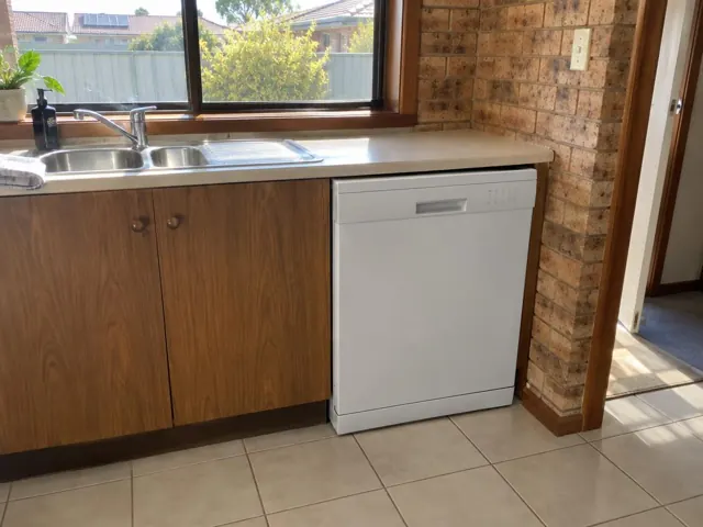 White dishwasher fitted under a kitchen benchtop beside a stainless sink and timber cabinets in Beresfield