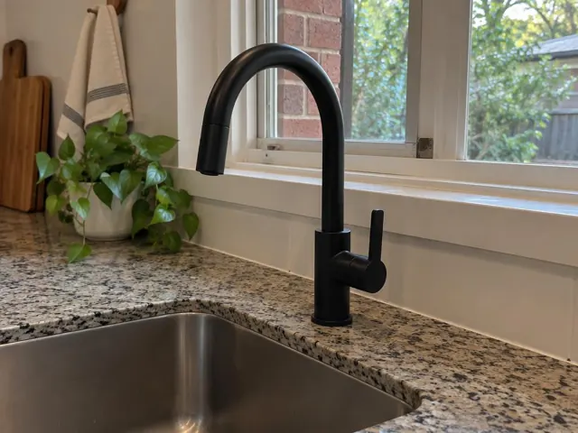 Black kitchen mixer tap beside a stainless sink under a window with speckled stone benchtop in Buttai